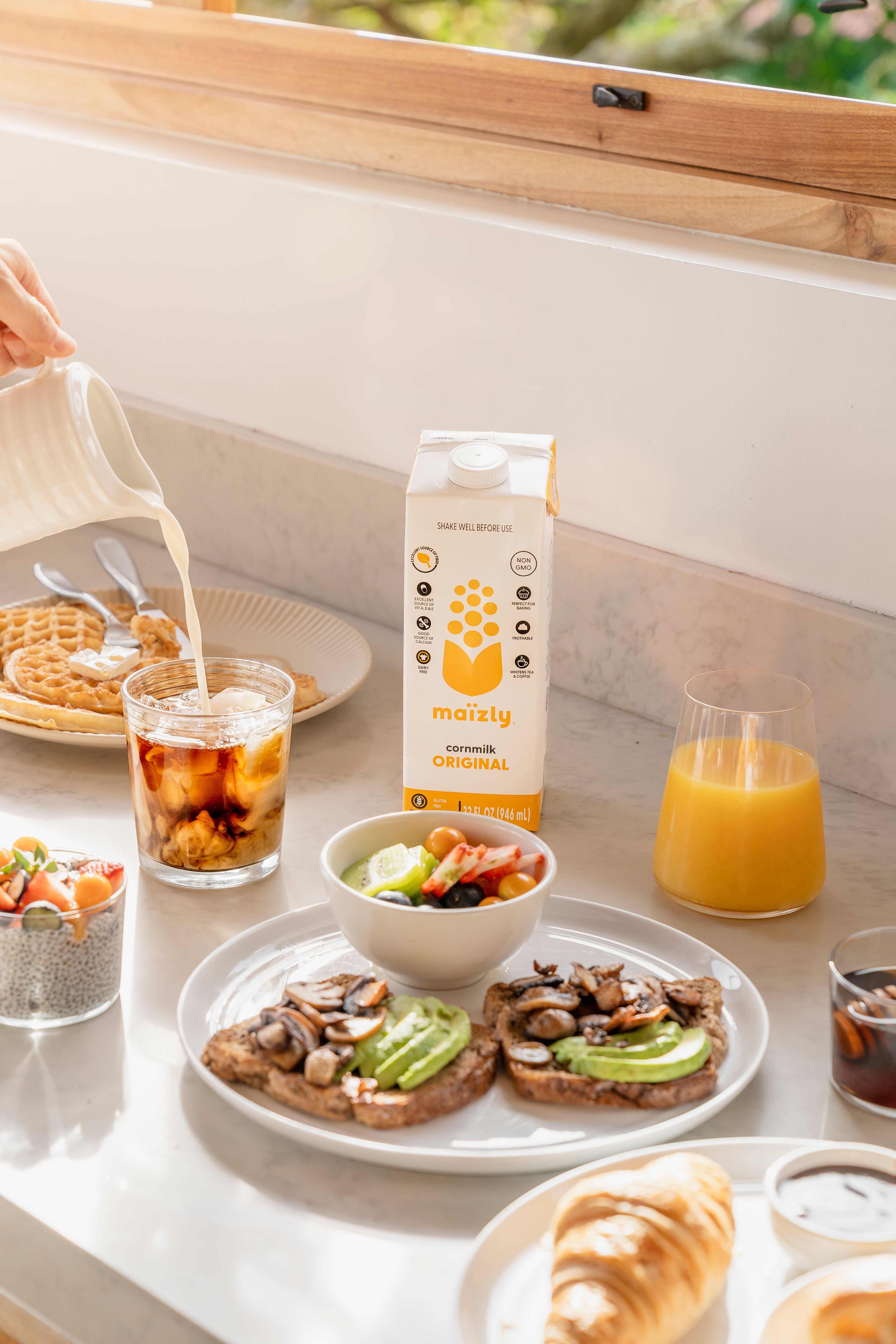 A sunny breakfast spread featuring Maïzly Original corn milk being poured into a glass of iced coffee, surrounded by avocado toast, fresh fruit bowls, chia pudding, waffles, and a glass of orange juice.