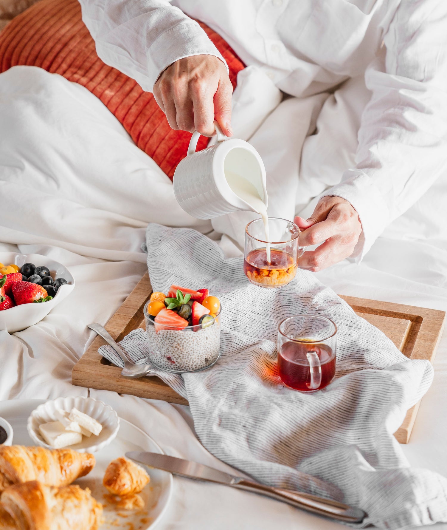 A cozy breakfast-in-bed scene featuring a person pouring Maïzly corn milk into a glass of tea, accompanied by a tray with chia pudding topped with fresh fruit, a bowl of mixed berries, and flaky croissants.