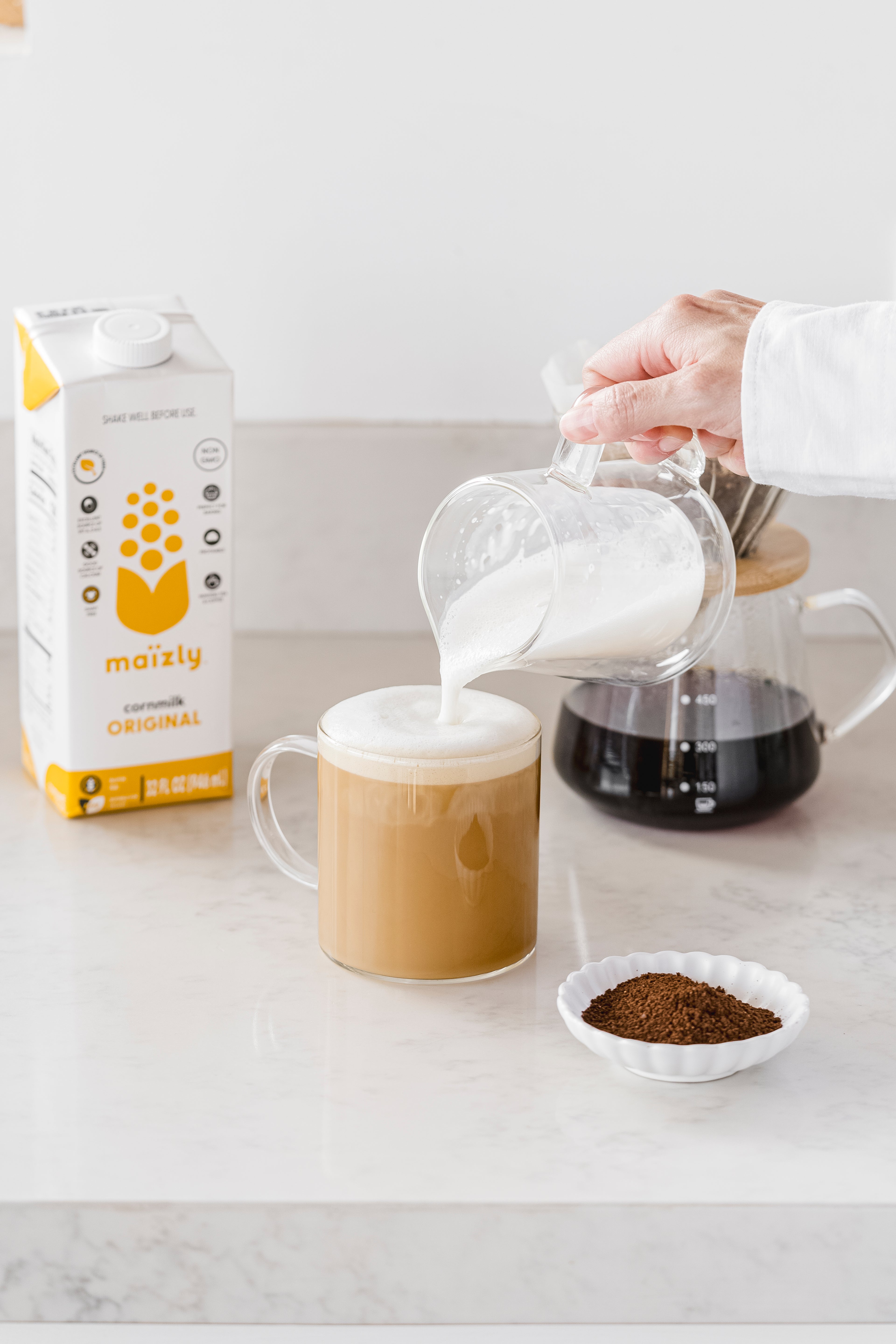 A person pouring Maïzly Original corn milk into a frothy coffee mug, with a carton of Maïzly, a French press coffee maker, and a small dish of ground coffee on a clean countertop.