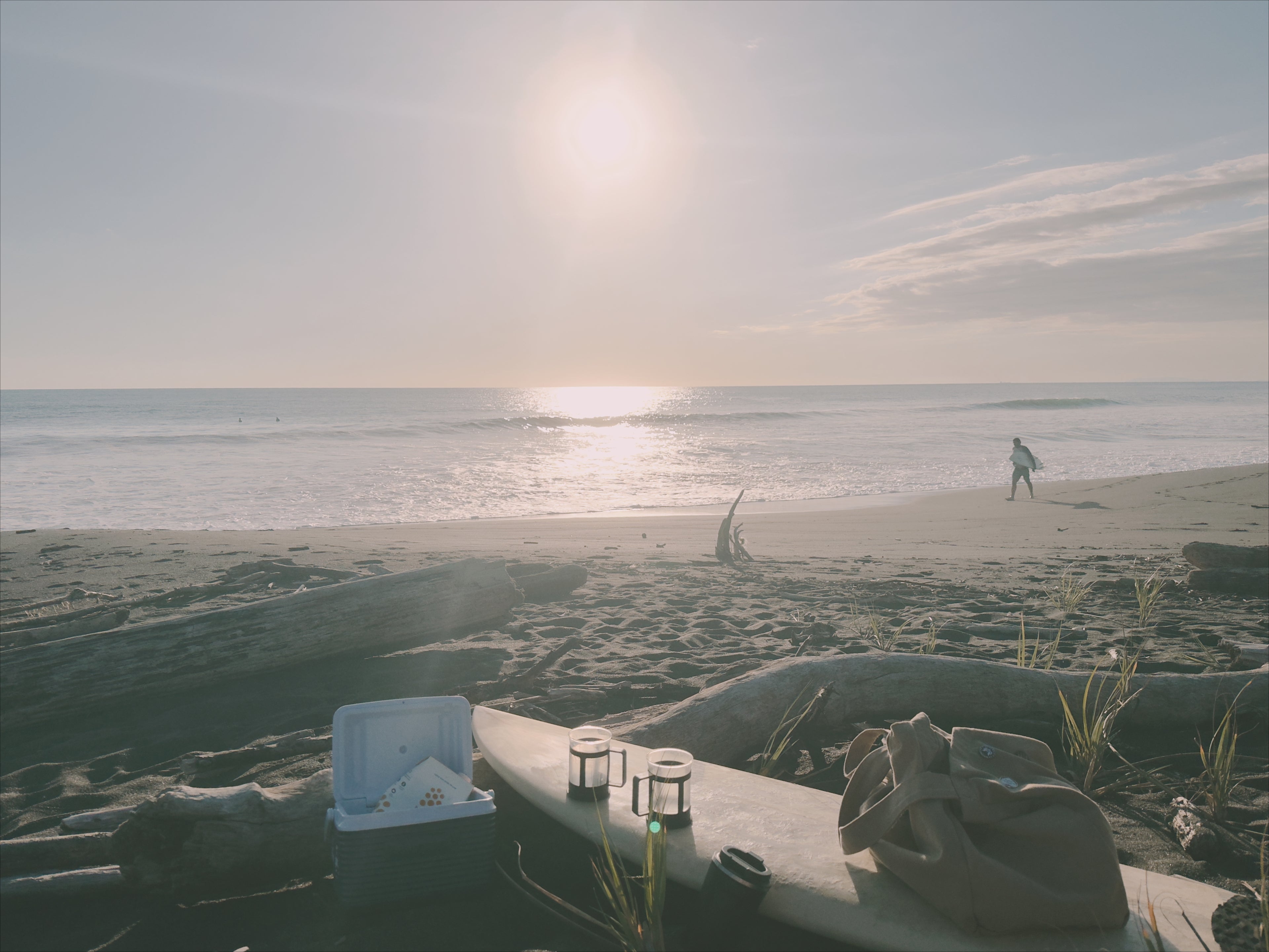 A serene beach scene at sunrise with a surfboard, a cooler containing Maïzly corn milk, and a French press coffee maker in the foreground, while a surfer walks along the shoreline in the distance.