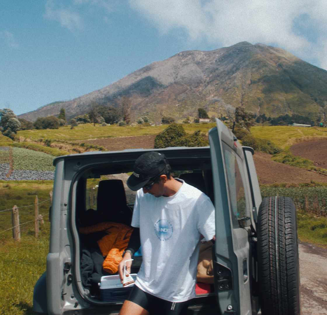 A man standing by the open trunk of a car, holding a Maïzly chocolate cornmilk, with a scenic backdrop of lush green hills and a mountain under a bright blue sky.