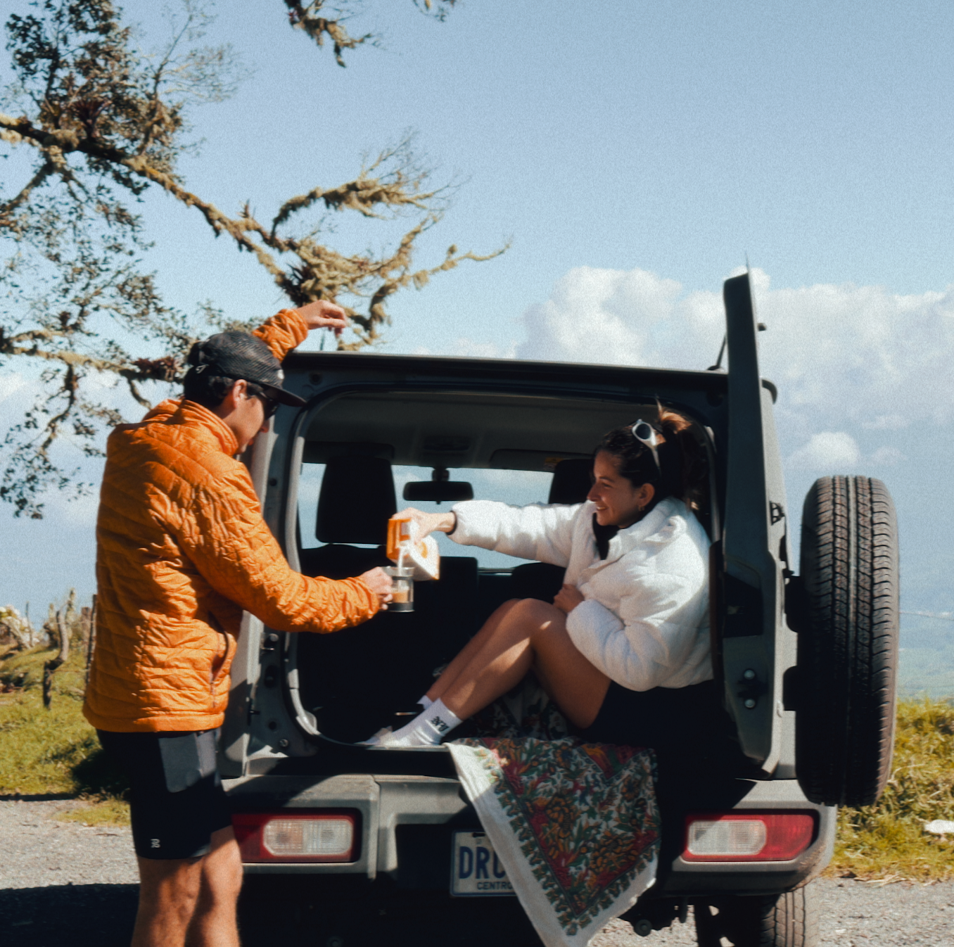 A man and woman enjoying a moment by the open trunk of a car. The man pours Maïzly into a glass while the woman, seated on a colorful blanket in the trunk, smiles. A scenic backdrop of trees and sky completes the outdoor vibe.