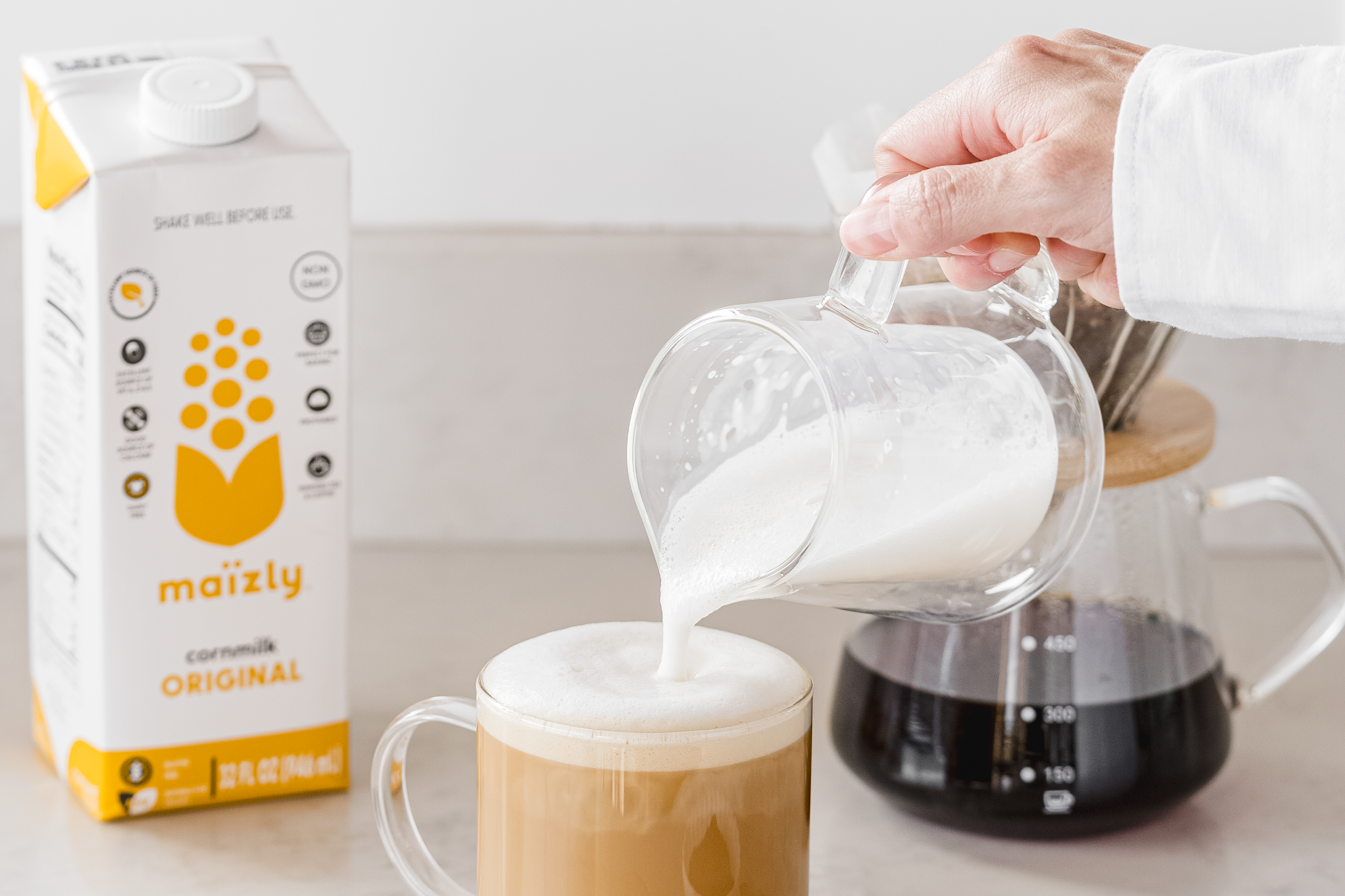 A hand pouring Maïzly Original corn milk into a frothy coffee mug, with a carton of Maïzly in the background next to a glass coffee carafe filled with freshly brewed coffee.